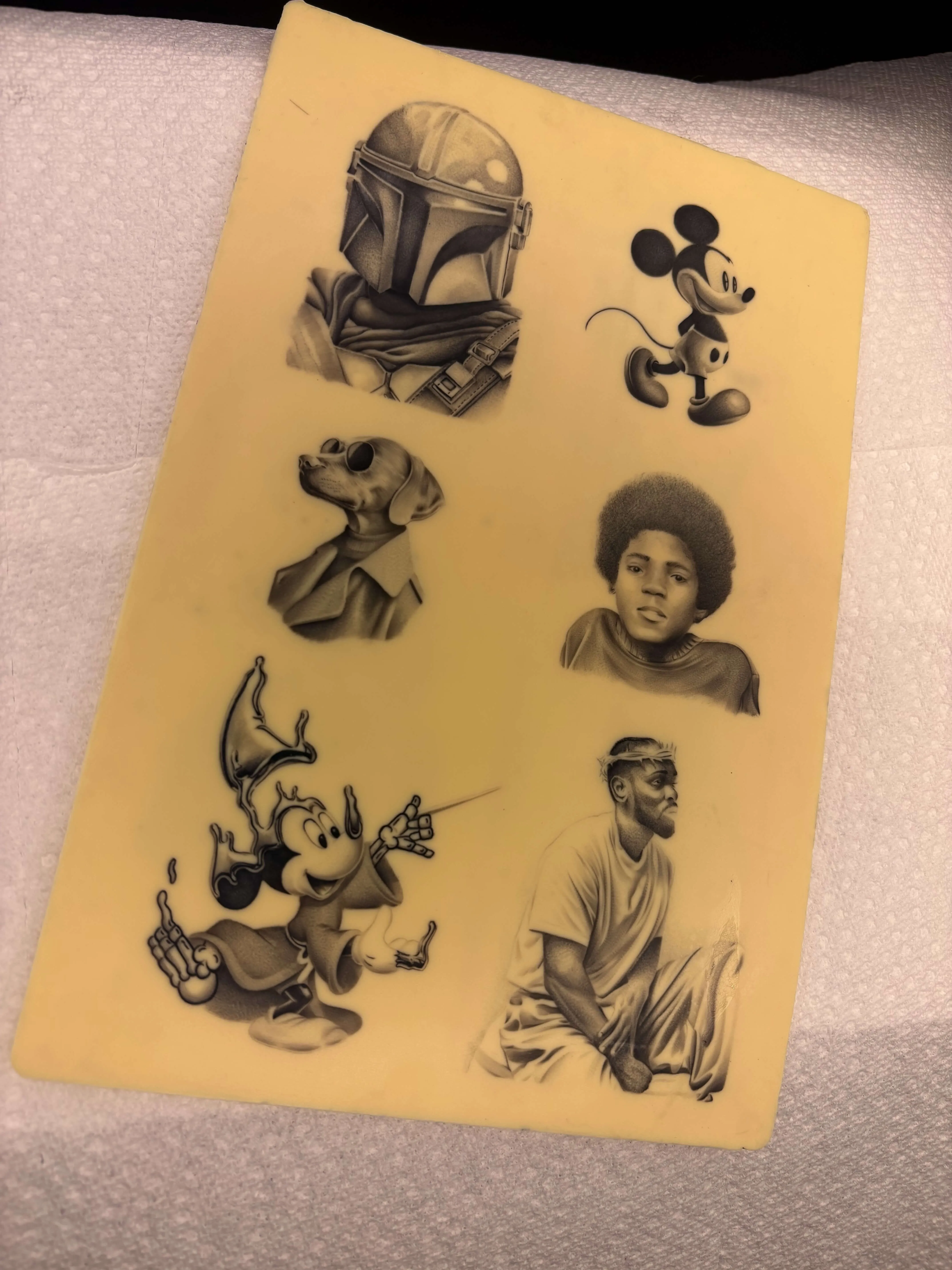 Synthetic tattoo practice skin sheet displaying black and grey realism flash designs including portraits and pop culture characters